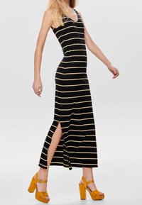 Abito maxi a righe nere e gialle con silhouette aderente e spacco laterale, abbinato a sandali con tacco alto gialli e spessi.