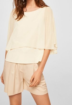 Kvinde iført en løs, cremefarvet lagdelt bluse med albuelange ærmer og højtaljede beige plisserede shorts.