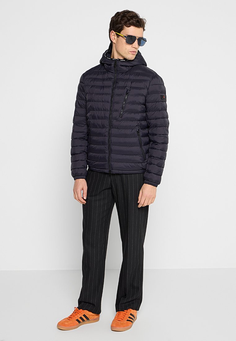 Giacca imbottita navy con trapuntatura orizzontale, chiusura con zip e cappuccio; abbinata a pantaloni neri a righe verticali e sneakers arancioni.