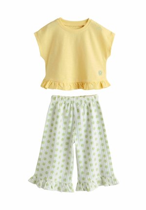 Žltý top s krátkym rukávom, nariaseným lemom a malou slnečnou grafikou, skombinovaný s bielymi nohavicami širokého strihu s zelenými slnečnými vzormi a nariasenými manžetami.