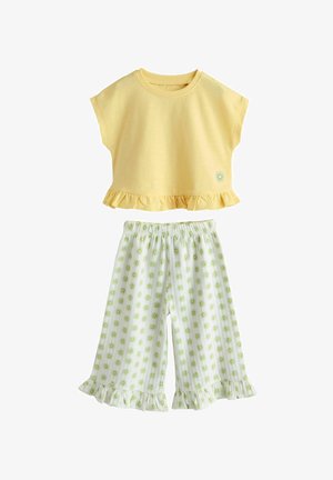 Žuti top kratkih rukava s naboranim rubom i malim grafičkim prikazom sunca, uparena s bijelim hlačama širokih nogavica koje imaju zelene uzorke sunca i naborane manžete.