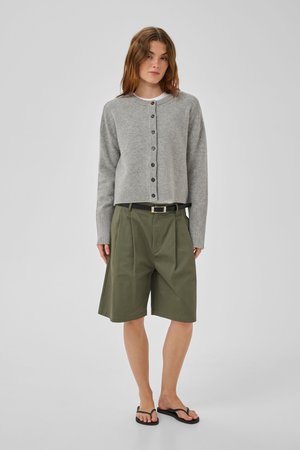Žena stojaca oblečená v sivom zapínacom cardigane, široké olivovo zelené šortky po kolená s čiernym opaskom a čierne žabky na jednofarebnom pozadí.