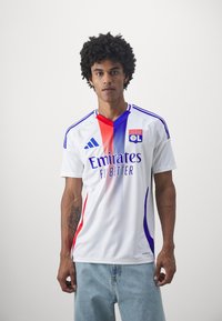 Bílý fotbalový dres s červenými a modrými akcenty, s logem Adidas, logem "Emirates" a znakem klubu Lyon. Krátké rukávy, design s kulatým výstřihem.