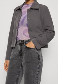 Giacca grigia corta con colletto e quattro bottoni, abbinata a una maglietta trasparente di un delicato viola e a jeans neri in denim a vita alta.