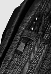 Zwarte rugzak met ritssluiting, voorzien van gestructureerde stof, meerdere ritsen en trekkerlipjes met logodetails. Close-up weergave gericht op het beslag.
