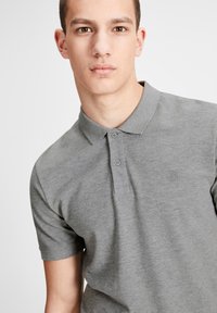 Polo shirt gris en tissu doux, avec col, patte de boutonnage à deux boutons et un petit logo sur la poitrine gauche. Manches courtes.