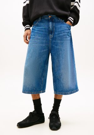 Persona che indossa pantaloncini blu in denim larghi fino al ginocchio, calzini neri alla caviglia, scarpe da ginnastica nere e un maglione nero con polsini a righe bianche.