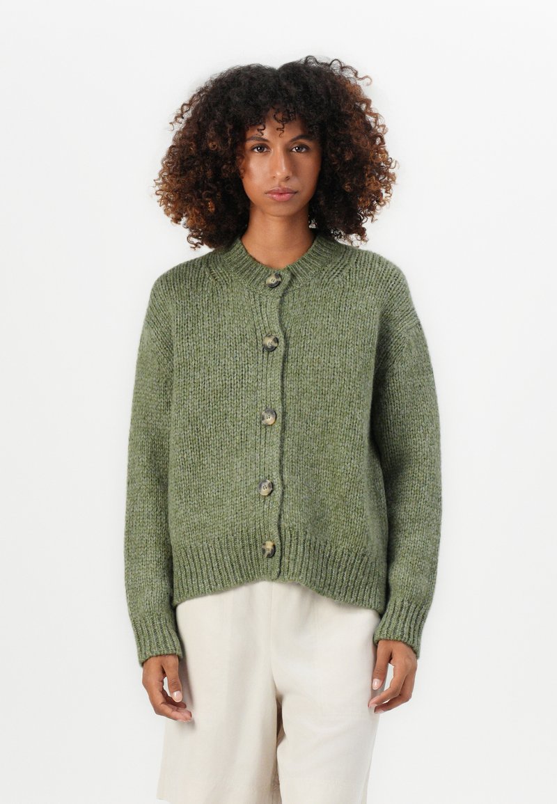 Giovane con capelli ricci indossa un cardigan verde oliva spesso con bottoni e pantaloni beige larghi, in piedi di fronte a uno sfondo bianco semplice.
