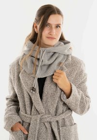 Jonge vrouw in een gestructureerde beige jas en een dikke lichtgrijze gebreide sjaal, staand met een hand in haar zak en de andere hand die de sjaal vasthoudt.
