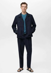 Marineblaues Leinenjackett mit zwei Brusttaschen, kombiniert mit einem türkisfarbenen Pullover, marineblauen Hosen und schwarzen Sandalen, stehend vor einem weißen Hintergrund.