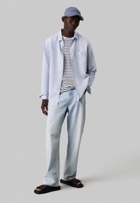 Camicia a maniche lunghe di colore azzurro chiaro sopra una maglietta a righe, abbinata a jeans in denim chiaro e sandali neri, completata da un cappellino da baseball lavanda.
