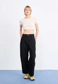 T-shirt beige clair de coupe ajustée avec des manches courtes ; pantalon noir ample avec un devant à boutons ; baskets jaunes avec des accents noirs ; fond uni.