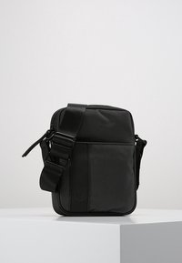 Malá černá crossbody taška s nastavitelným popruhem, texturovanou látkou a přední kapsou, stojící na bílém podstavci na jednobarevném pozadí.