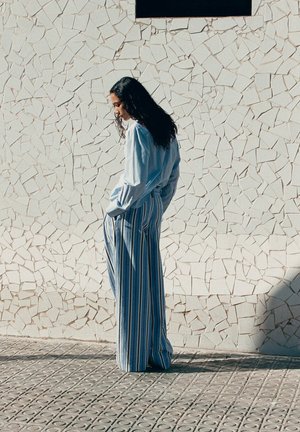 Mujer con cabello largo y oscuro, que lleva una blusa azul clara y pantalones anchos con rayas, de pie con las manos en los bolsillos frente a una pared blanca con textura.