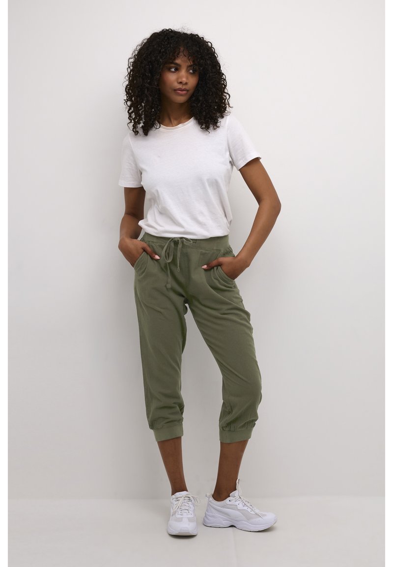 Femme aux cheveux bouclés portant un t-shirt blanc, un pantalon de jogging court vert olive et des baskets blanches, debout les mains dans les poches devant un fond uni.