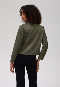 Krátká olivově zelená semišová bunda s kulatým límcem a dlouhými rukávy, kombinovaná s úzkými černými kalhotami. Hladká textura, minimalistický design.