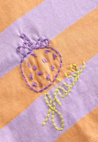 Un tissu rayé violet et orange présente un motif de fraise brodé en fils violet et jaune avec des points de décoration.