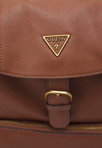 Sac à dos en cuir texturé marron avec quincaillerie dorée, doté d'un logo triangulaire, d'une sangle réglable et de détails de poche zippée.