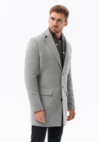 Cappotto grigio in lana con revers a notched, bottoni frontali e due tasche laterali, indossato sopra un maglione verde a trama e una sciarpa a quadri.