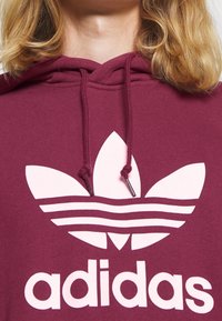 Maroon hoodie med en stor vit Adidas trefoil-logotyp och tre horisontella ränder nedanför. Dragband fästa vid huvan.