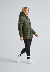 Doudoune vert olive avec capuche, fermeture éclair, poches latérales et logo sur l'épaule, portée avec des leggings noirs et des baskets blanches.