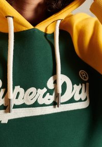 Felpa con cappuccio verde e gialla con grande logo ricamato "SuperDry" bianco, con spacchetti bianchi e una consistenza morbida.