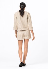 Beige texturerad stickad set som innehåller en kort jacka med vikta ärmar och matchande shorts. Mjuk tyg och fransade kanter. Svarta slip-on skor.