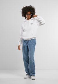 Felpa bianca con tasca a marsupio, caratterizzata da un piccolo dettaglio grafico. Abbinata a jeans azzurri chiari e larghi e scarpe da ginnastica bianche.