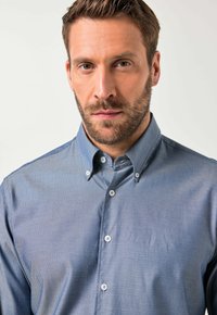 Uomo con barba che indossa una camicia button-up azzurra chiara a fantasia con colletto bottone, che guarda direttamente nella fotocamera, sfondo neutro.