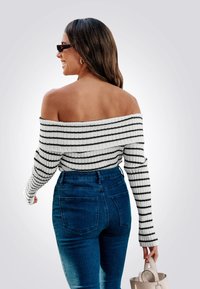 Maglione a coste off-the-shoulder bianco con strisce nere, abbinato a jeans in denim blu a vita alta; modella che tiene una borsa beige.