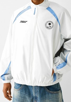 Giacca bianca con cerniera, dettagli azzurri, testo nero "TRAY" sul petto sinistro, logo circolare con rosa e testo sul petto destro, indossata sopra un paio di jeans.