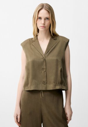 Donna dai capelli biondi lisci indossa una camicia smanicata verde oliva con bottoni e pantaloni larghi abbinati su sfondo bianco neutro.