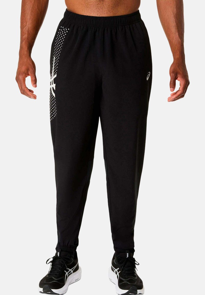 Homme portant un pantalon de jogging athlétique noir avec un logo blanc et des points sur la cuisse gauche, des chaussures de course noires avec des semelles blanches sur fond blanc.