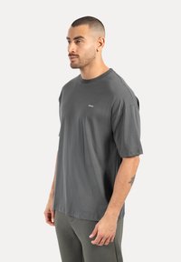 Mand med kort hår og skæg, der modellen en mørkegrå oversized T-shirt og bukser, står sidelæns mod en ensfarvet baggrund.