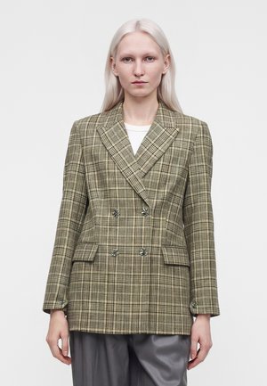 Femme aux longs cheveux blonds platine porte un blazer croisé à carreaux verts et un pantalon gris, debout devant un fond clair uni.