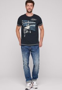 Camiseta azul marino con texto e imágenes impresas, combinada con jeans de denim azul claro. Zapatillas con acentos rojos y azules completan el look.