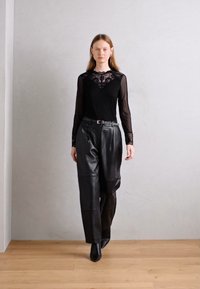 Haut à manches longues noir avec des détails en dentelle, associé à un pantalon en cuir noir taille haute et des bottines noires à talons pointus, sur un sol en bois clair.