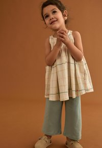 Weiß karierte Tunika mit bunten Blumenakzenten, kombiniert mit hellblauen strukturierten Hosen und beigen Slippers. Der Hintergrund ist braun.
