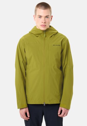 Junger Mann trägt olivgrüne wasserdichte Kapuzenjacke mit Frontreißverschluss und schwarze Hose, steht vor weißem Hintergrund.