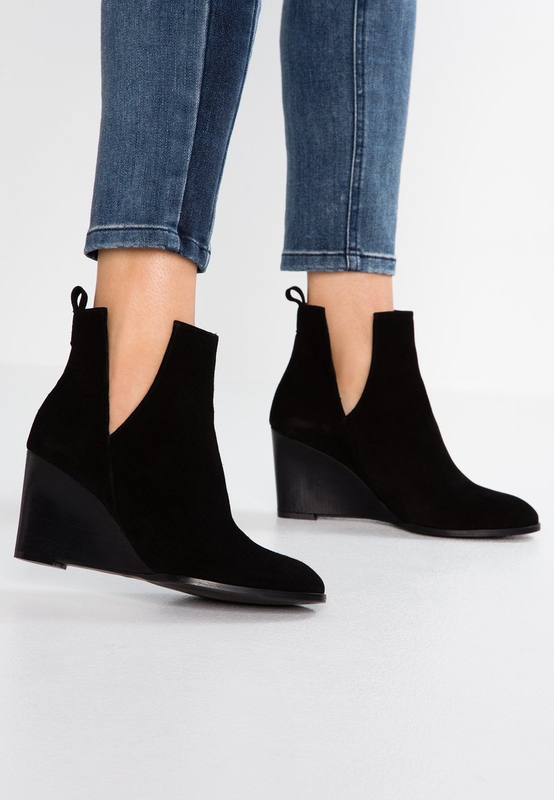 Personne portant des bottines compensées noires en daim et un jean skinny bleu retroussé sur un sol et un fond blanc.