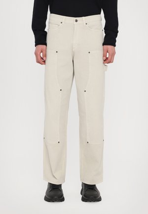 Light beige cargo pants with knee seams, dual side pockets, and black hardware. Worn with black shoes, featured against a plain background.