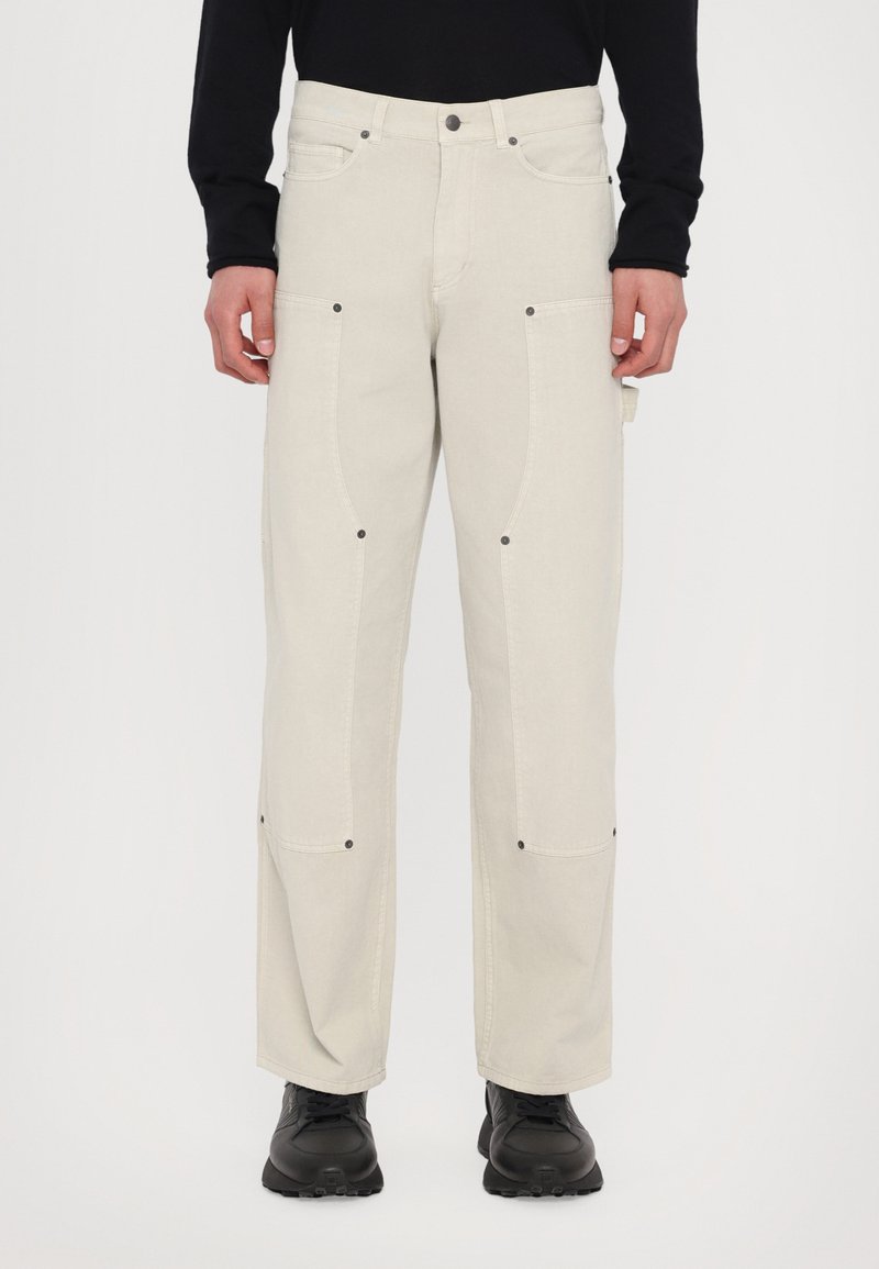 Light beige cargo pants with knee seams, dual side pockets, and black hardware. Worn with black shoes, featured against a plain background.