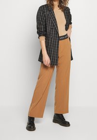 Blazer a scacchi neri con design doppiopetto, dolcevita aderente beige e pantaloni marroni a gamba larga; stivali neri con lacci.
