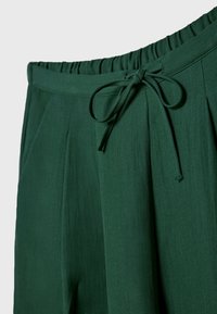 Groene broek met elastische tailleband en stoffen trekkoord dat in een strik is geknoopt, met zijnaadzakken, op een effen achtergrond.