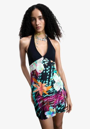 Femme aux cheveux bouclés portant une robe noire à col licou avec des motifs floraux vifs et abstraits en turquoise, rose et orange, accessoirisée d'un collier en argent.