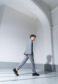 Szary garnitur z dopasowaną marynarką i zwężającymi się spodniami, w komplecie z błyszczącymi czarnymi butami. Gładka tekstura, chodząc po marmurowej podłodze z cieniem.