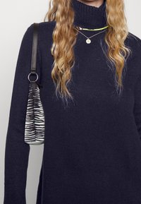 Marinblå polokragetröja med texturerad stickning, kompletterad med lager av halsband. En grå och svart zebramönstrad väska hänger från ett tunt band.