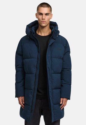 Junger Mann in einer navyblauen gepolsterten langen Winterjacke mit Kapuze und schwarzem Hemd, der vor einem schlichten hellen Hintergrund steht.