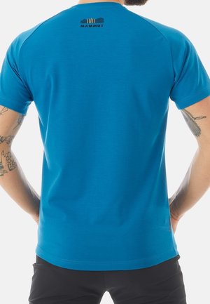 Muž v modrém tričku Mammut, na zádech u krku logo, viditelná tetovaná ramena a černé šortky na bílém pozadí.