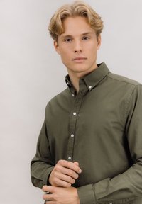 Olivengrønn knapp-skjorte med klassisk krage, med hvite knapper og lange ermer. Glatt tekstur, tettsittende stil, minimalistisk design.